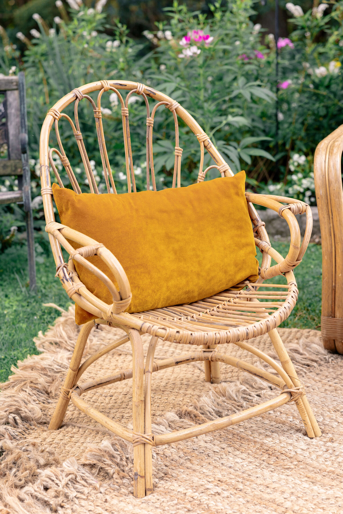 A bamboo chair with a mustard-colored cushion sits on a textured beige rug in an outdoor garden setting, surrounded by greenery and flowers.