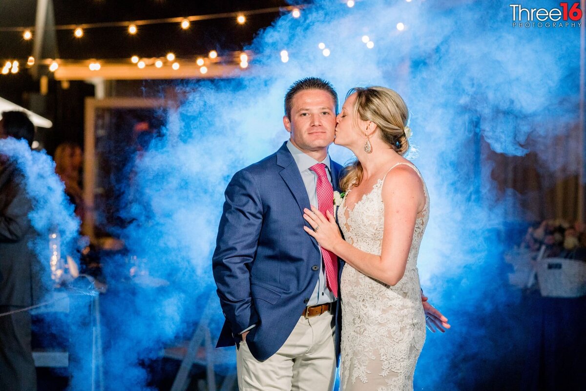 Bride kisses her Groom on the cheek during an evening photo shoot with blue mist behind them