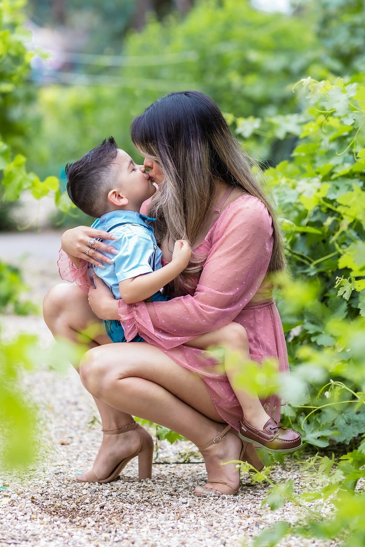 Mother is wearing a mauve long sleeve romper and is kissing her son while posed in in a vineyard