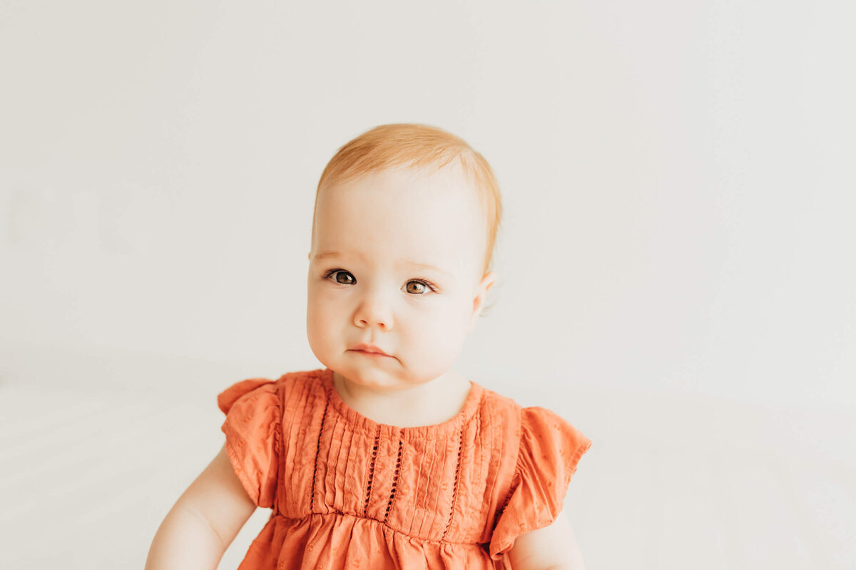 Baby H wearing a rust colored dress staring at camera in studio for Ally's Photography.