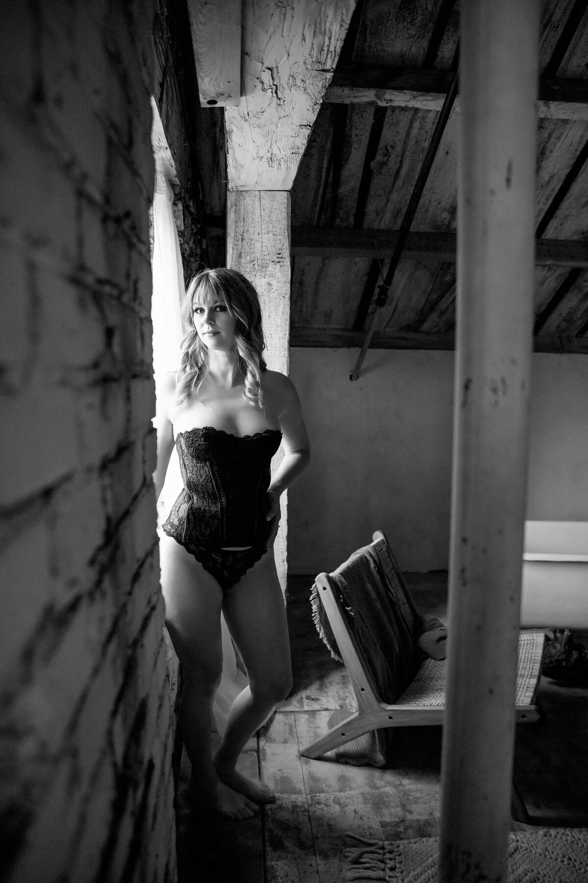 black and white photo of a lady wearing a black bodice and underwear standing by a window for her Mississauga boudoir photography session