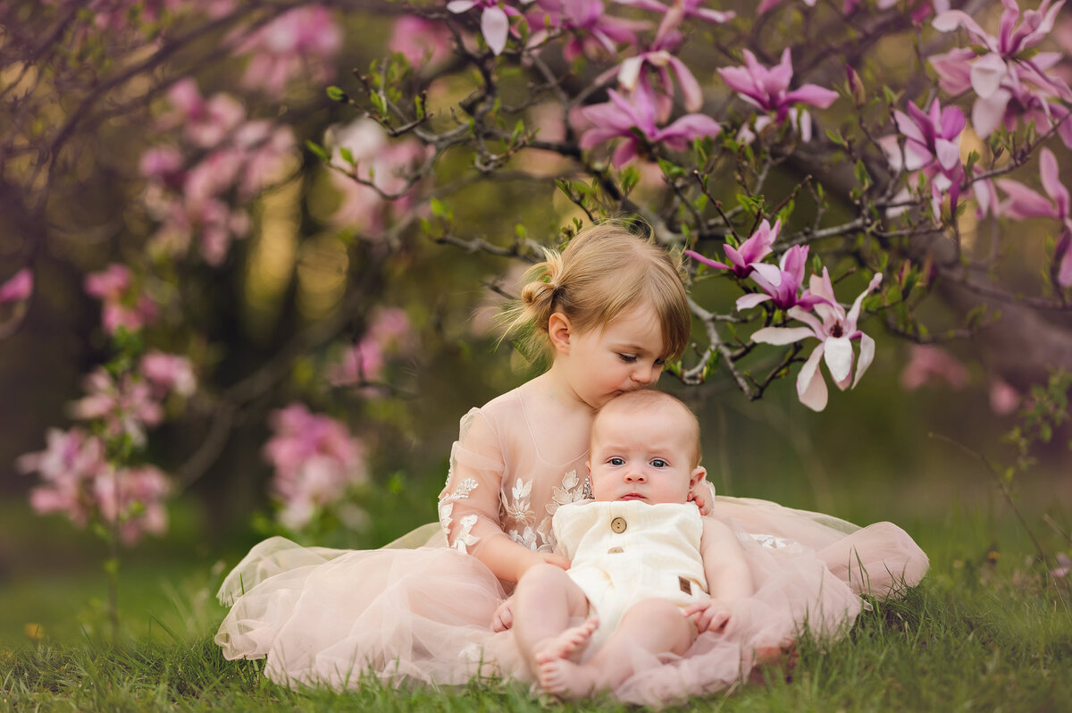 A young girl cradles her baby brother surrounded by pink magnolia blossoms.