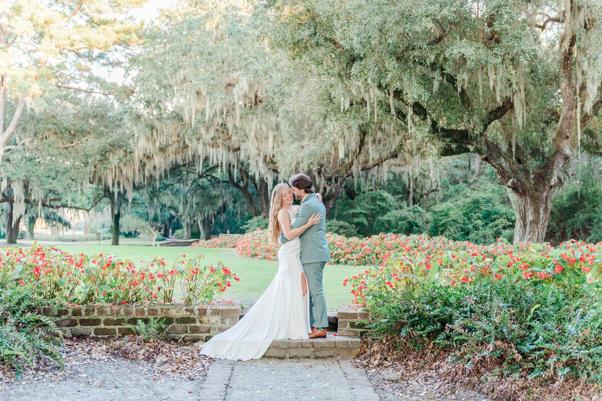 a bride and groom stand hugging each other in the middle of a beautiful garden with tall weeping Oak trees with swaying moss