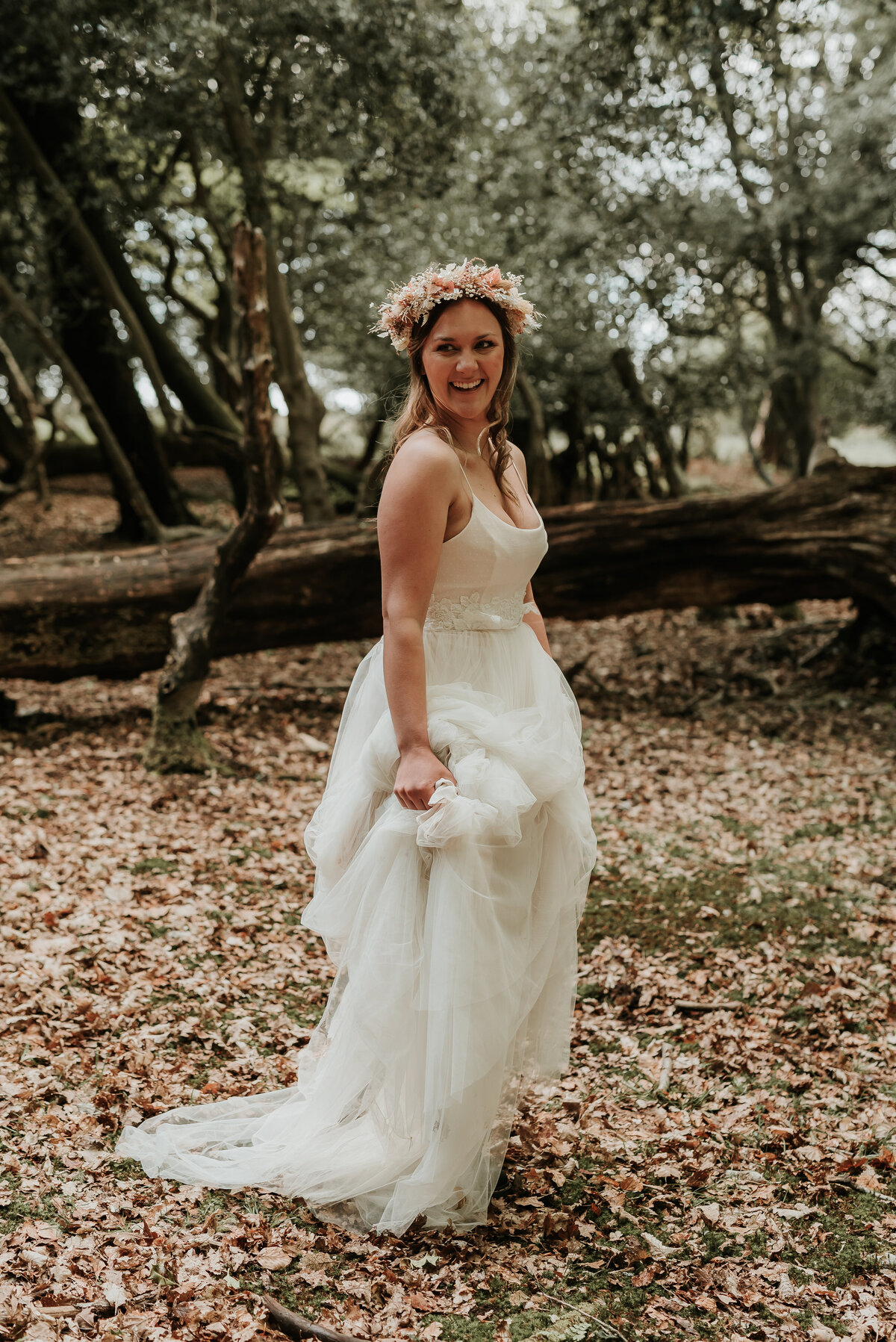 Bride wearing a dried floral crown gathers her dress as she walks through the woodlands in New Forest