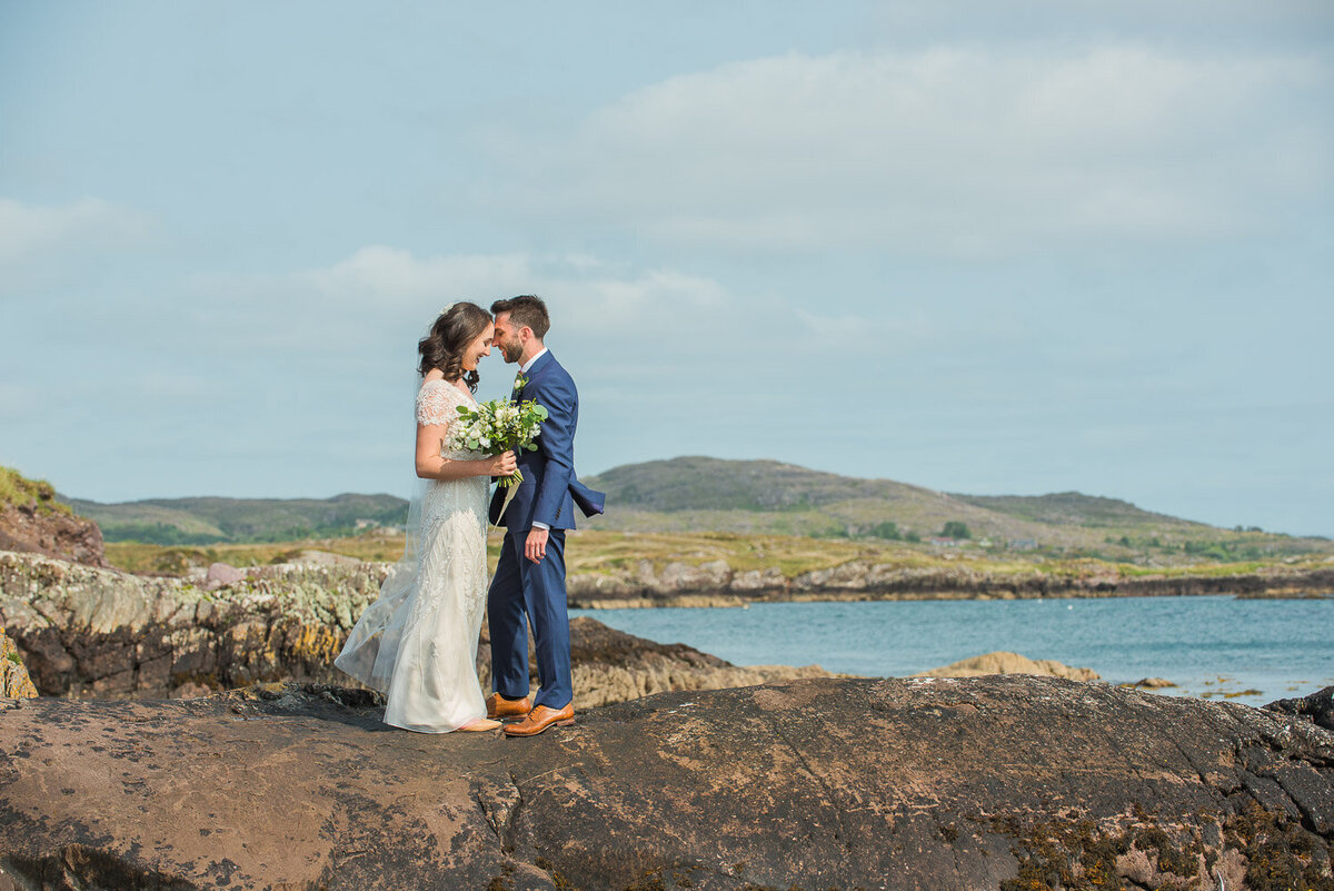 bride wearing a modified a-line, beaded wedding dress embracing her husband while standing on rocks overlooking Castlecove in Kerry