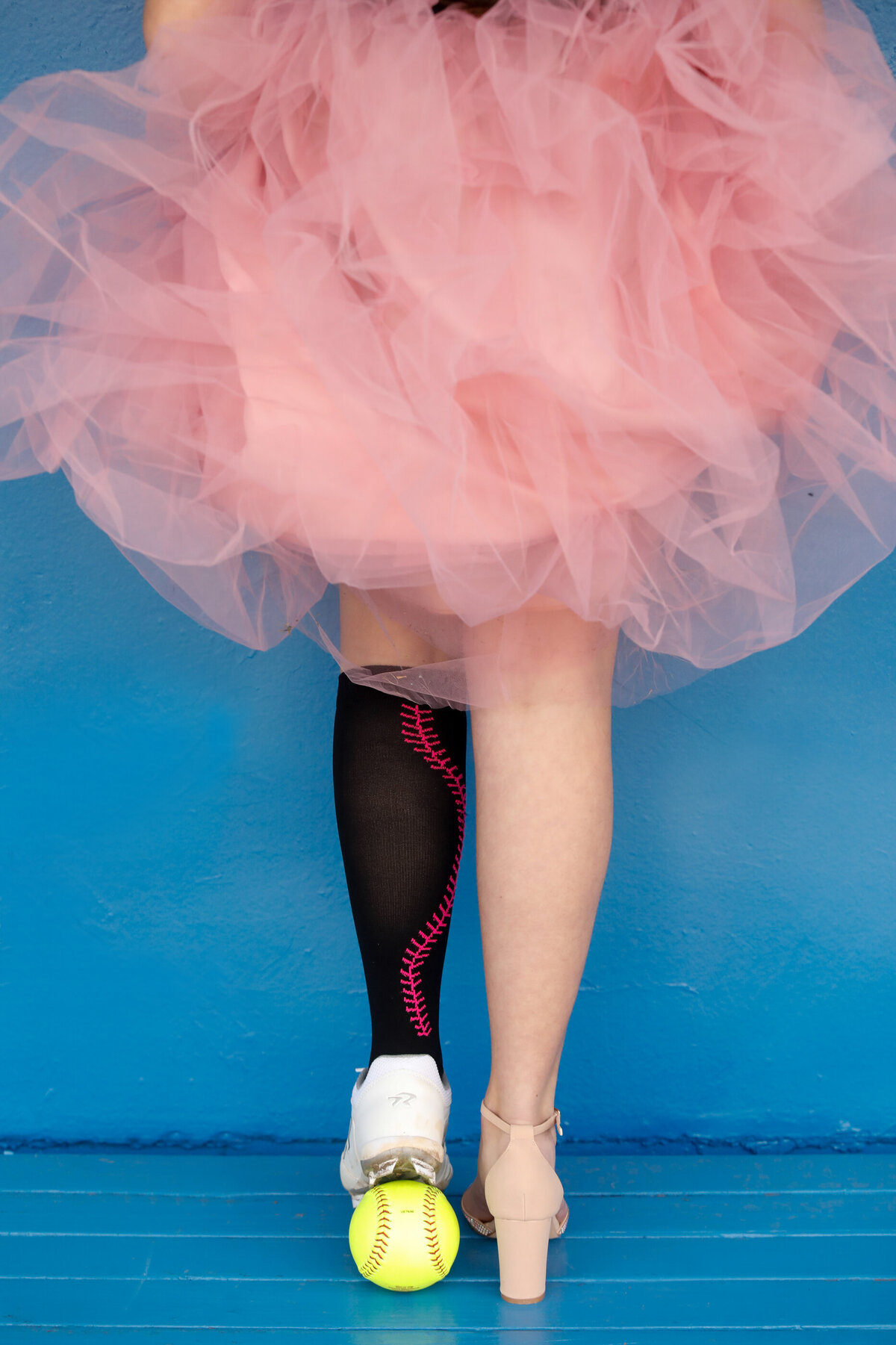 Detail shot of softball player's legs wearing a high heel on one foot and softball cleats and socks stepping on a ball while wearing a tutu Rockland County senior portrait photographer Milestone Images