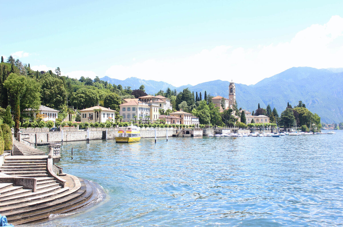 Scenic view of a lakeside town with elegant buildings and a church tower, surrounded by lush greenery and mountains in the background. A yellow boat is docked by the shore, and stone steps lead down to the water.