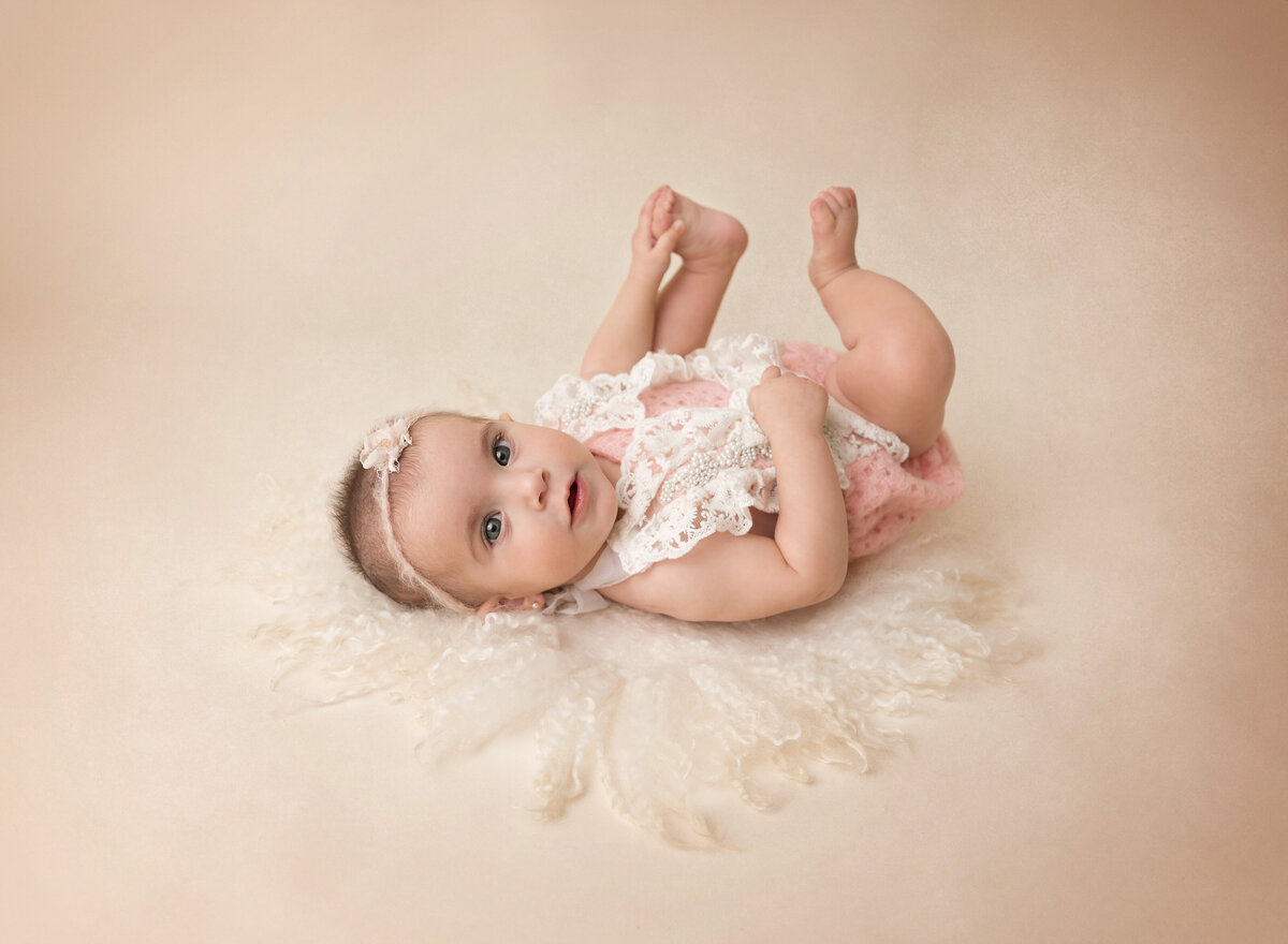 Baby girl lying on her back for a 6-month baby photo session. Baby is wearing a white lace onesie, holding her toes, and looking at the camera.