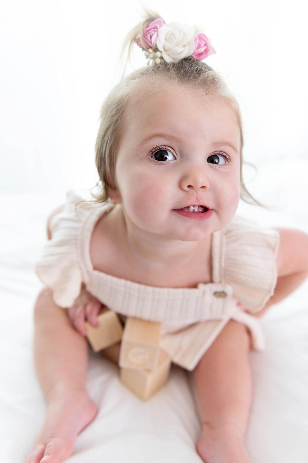 A toddler girl in a pink dress makes a silly face while sitting on a bed playing with wooden block