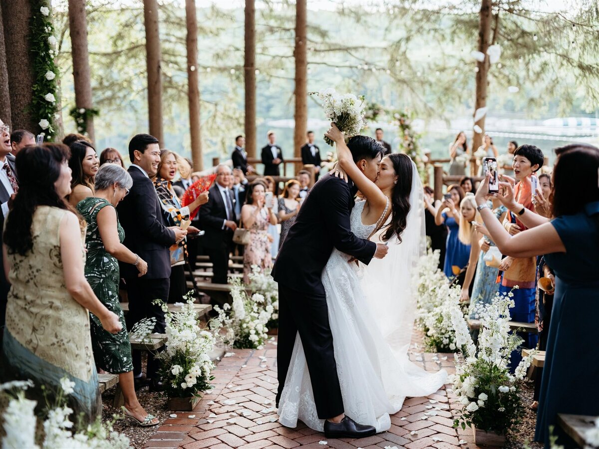 Bride and groom embrace for a kiss after wedding ceremony at the end of the aisle, surrounded by friends and family. Pine trees and lake in background at Cedar Lakes Estate wedding venue in Hudson Valley.