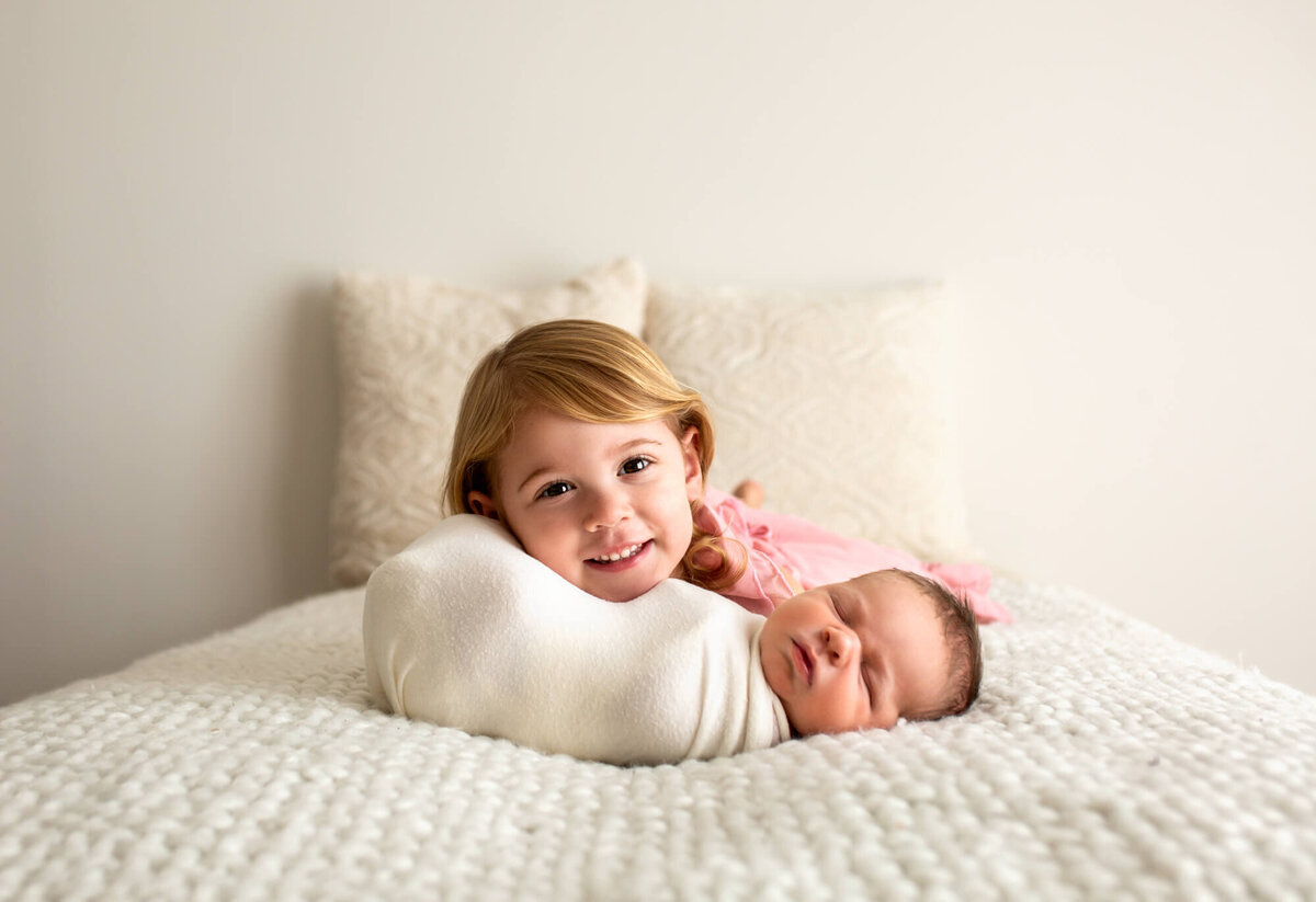 A big sissy snuggles her newborn brother and smiles for the camera during a portrait session with an Asheville Newborn Photographer