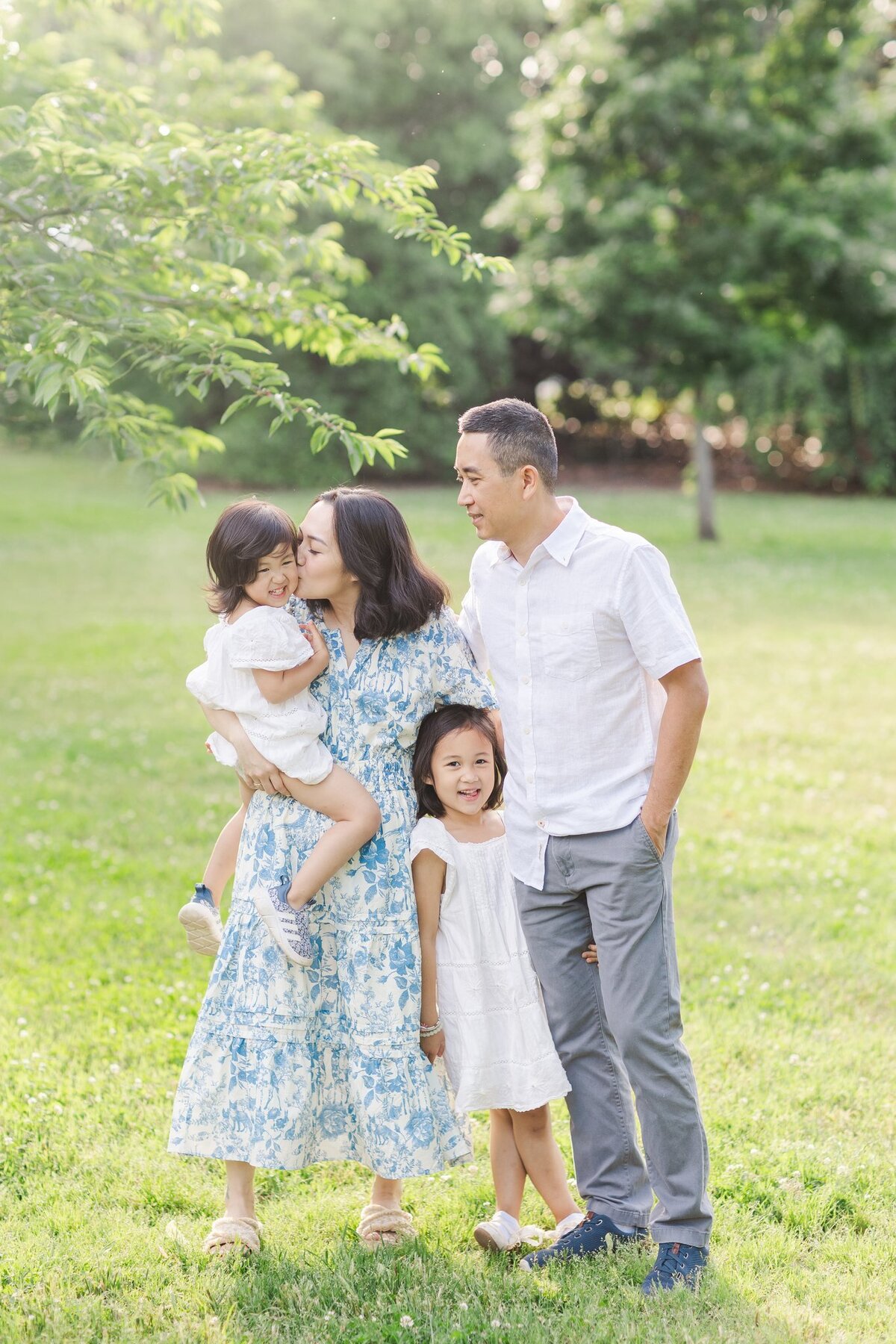 Mother kisses daughter's cheek while dad laughs at them and older daughter smiles at camera