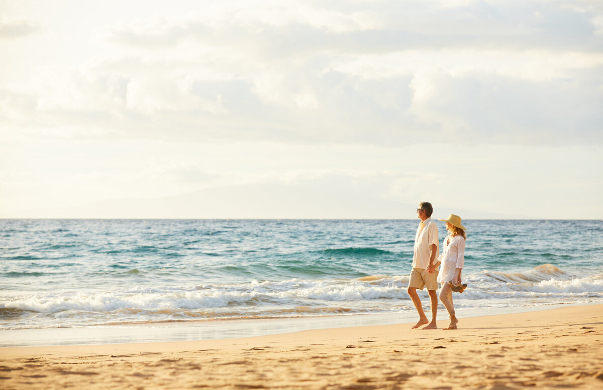 A couple walks hand in hand along a sandy beach with gentle waves in the background. They wear casual white clothing, and the woman has a sun hat. The sky is partly cloudy, creating a serene and romantic atmosphere.