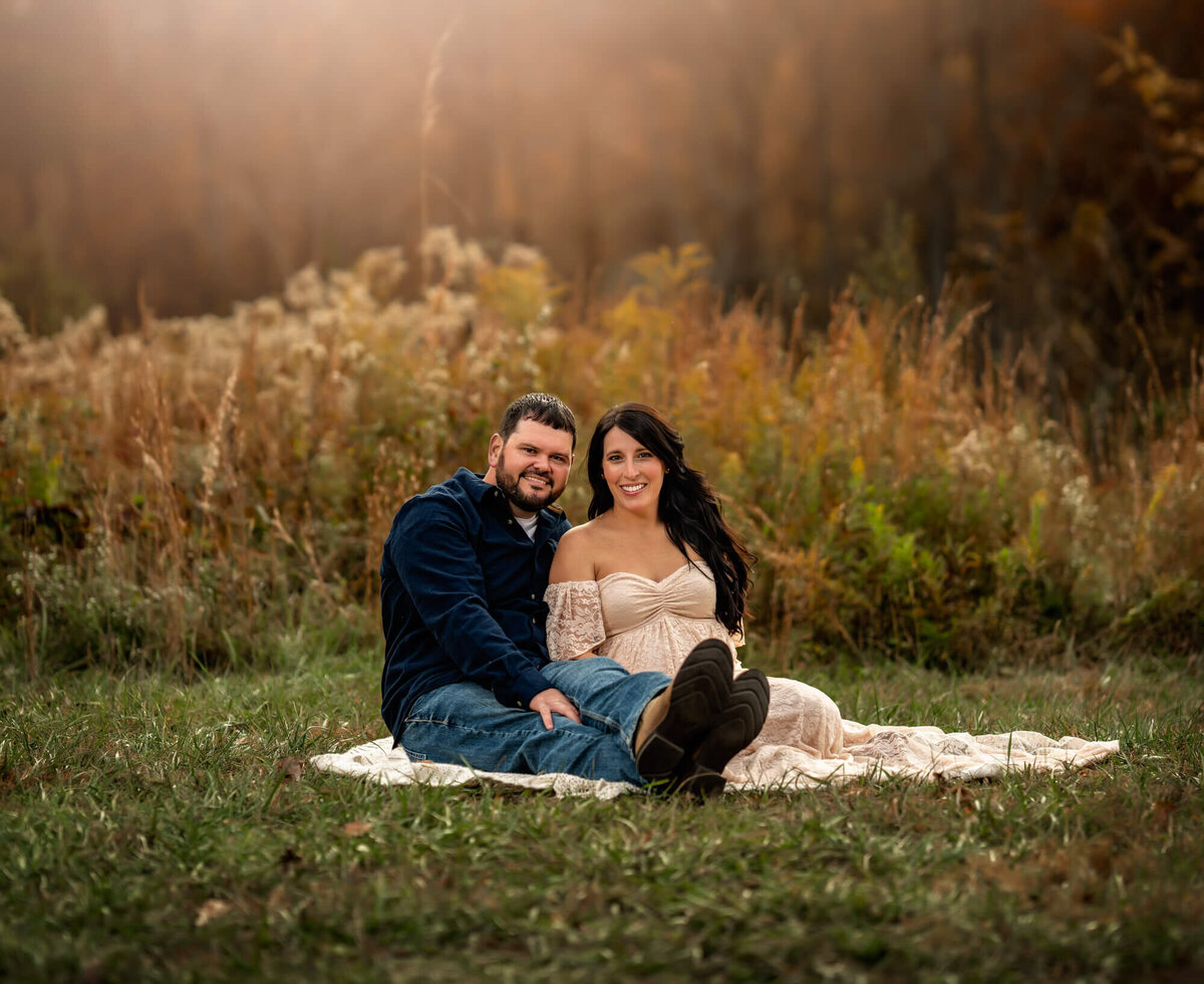 A mom and dad to be sit on a cream blanket with tall yellow grass behind them