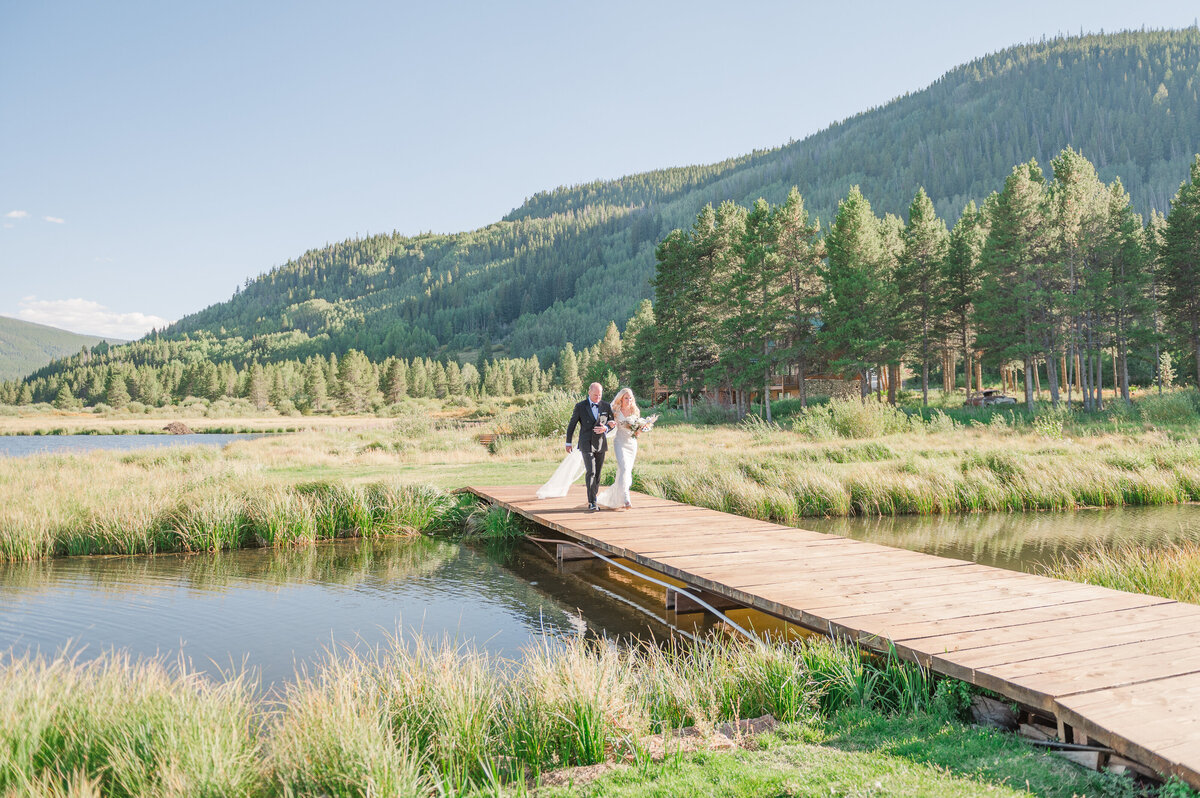 Dad and bride walking across a wooden foot bridge to an outdoor wedding ceremony in Camp Hale Colorado.
