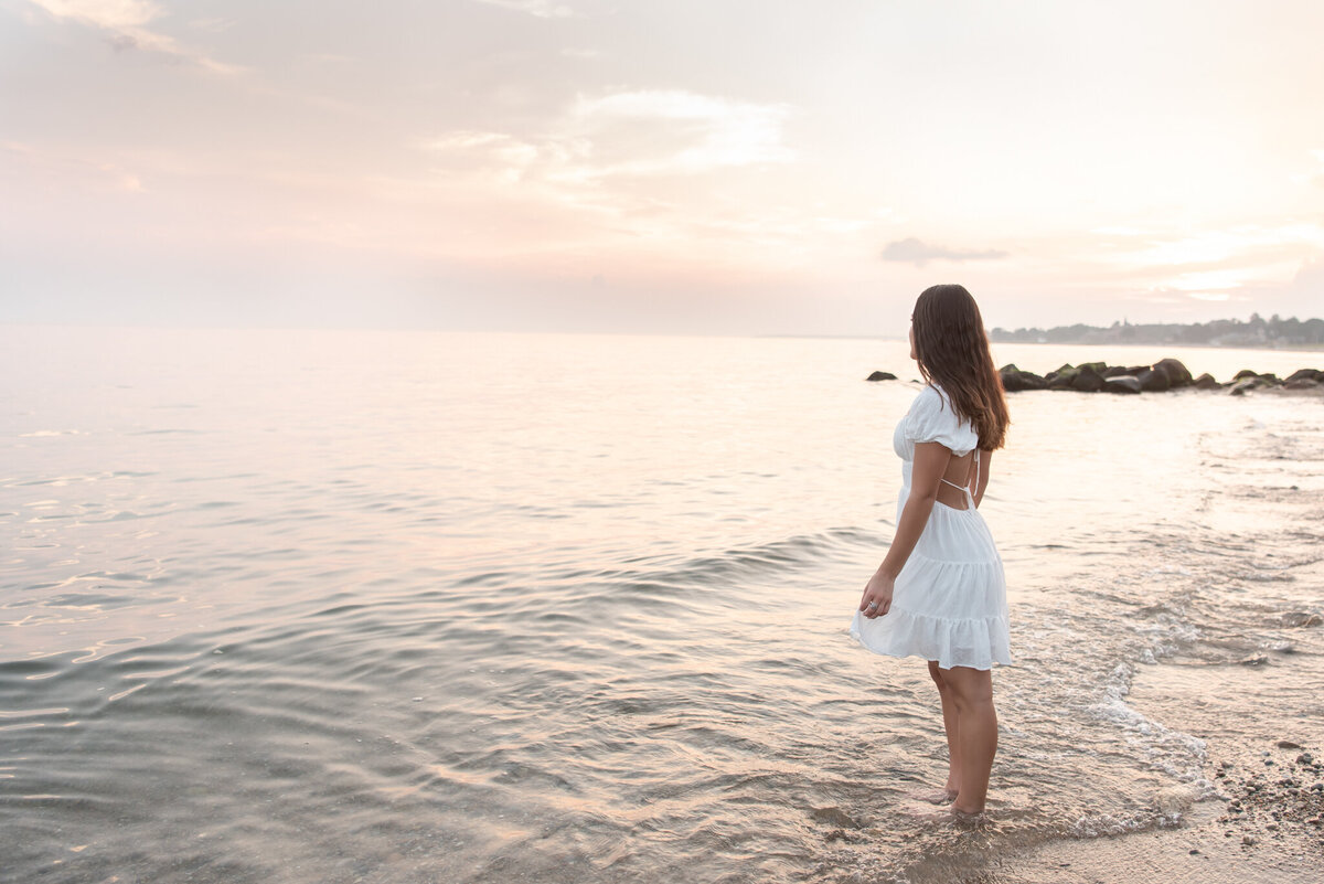 High school senior girl in white dress looking out over the ocean at sunset