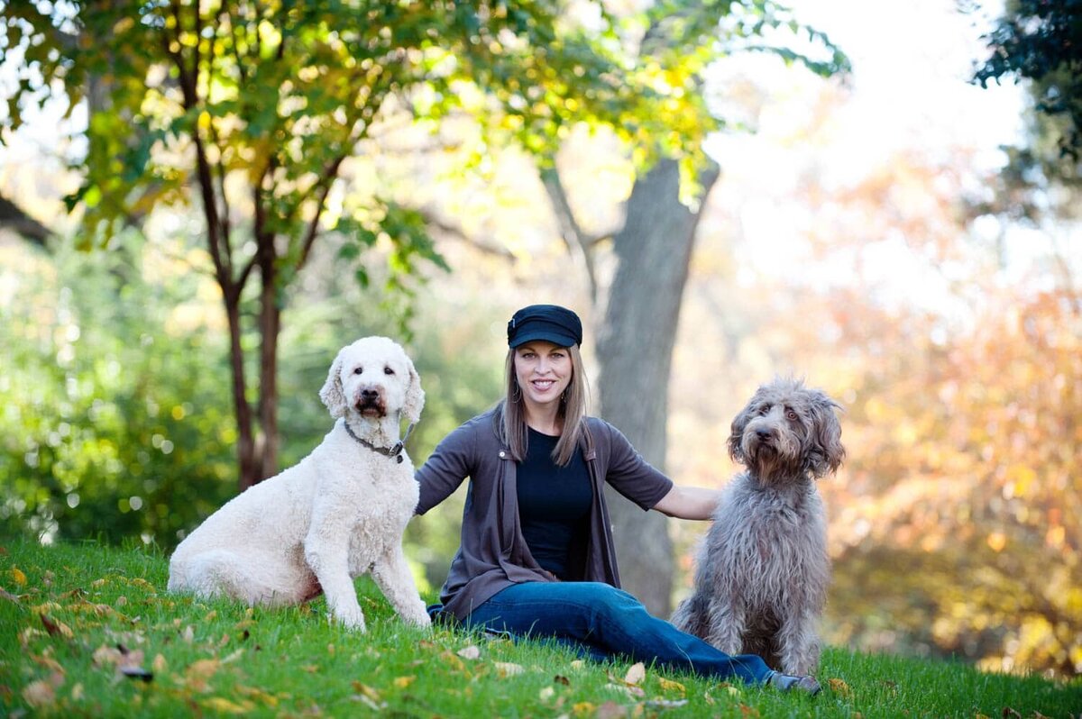 Woman wearing a hat sits and stays with her 2 shaggy dogs on each side of her in the park.
