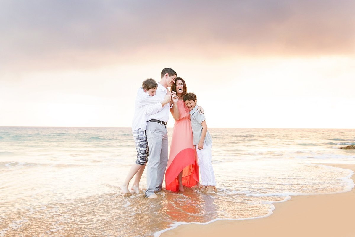 Mom in coral dress embraced by husband and teen boys during their family Maui portrait at the beach