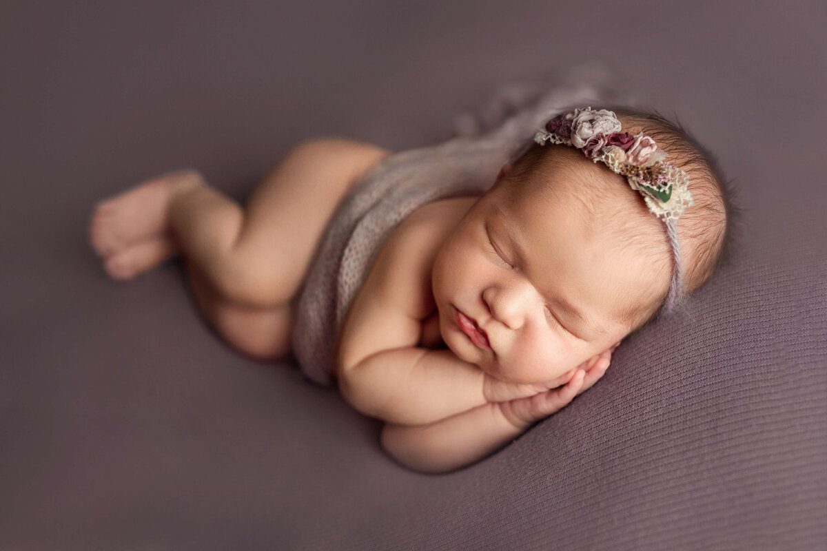 newborn abby laying on her side with a purple floral headband and purple wrap on a matching backdrop