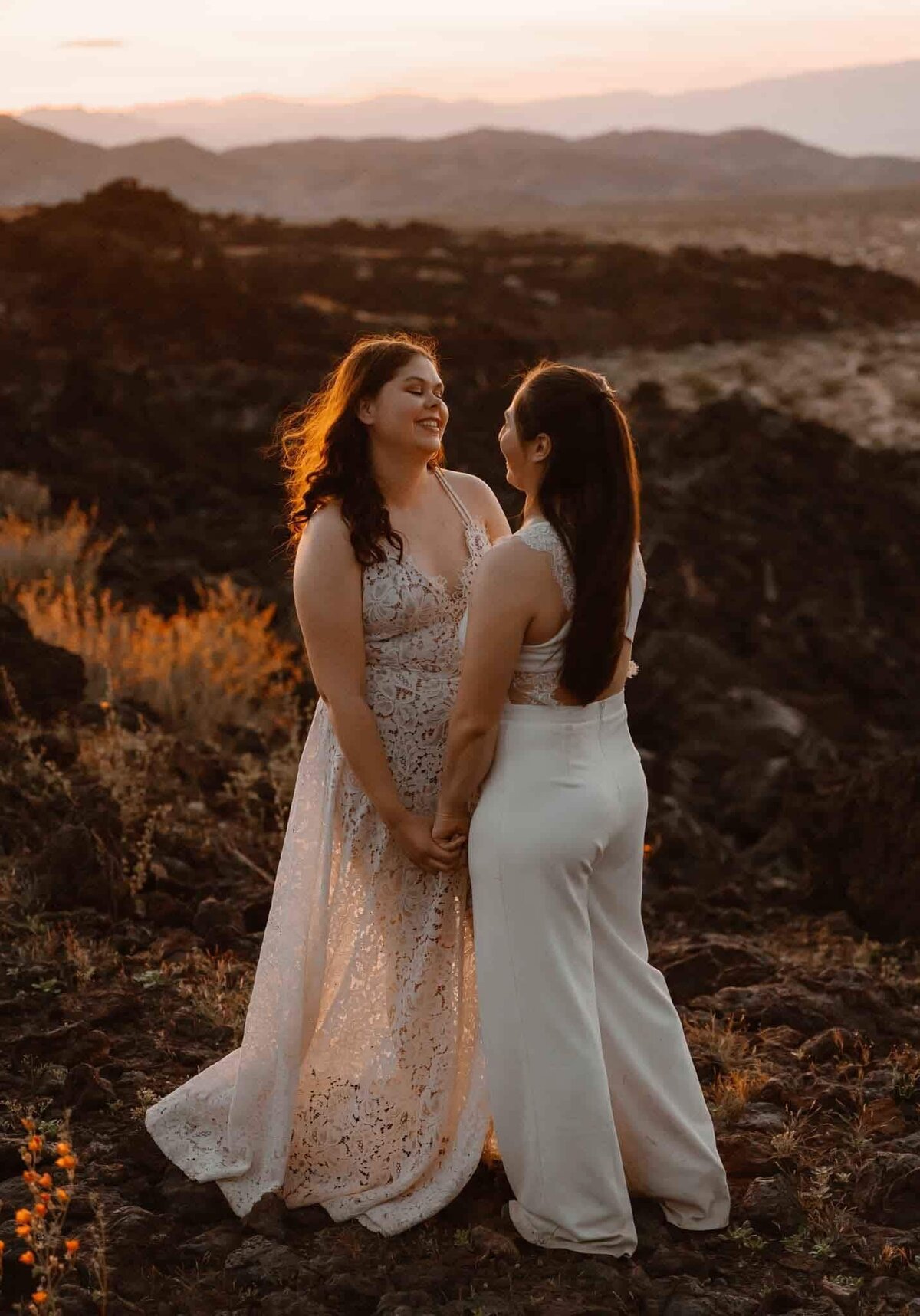A couple stands smiling at each other in a rocky, sunlit landscape, with dark lava rock and mountain silhouettes in the background.