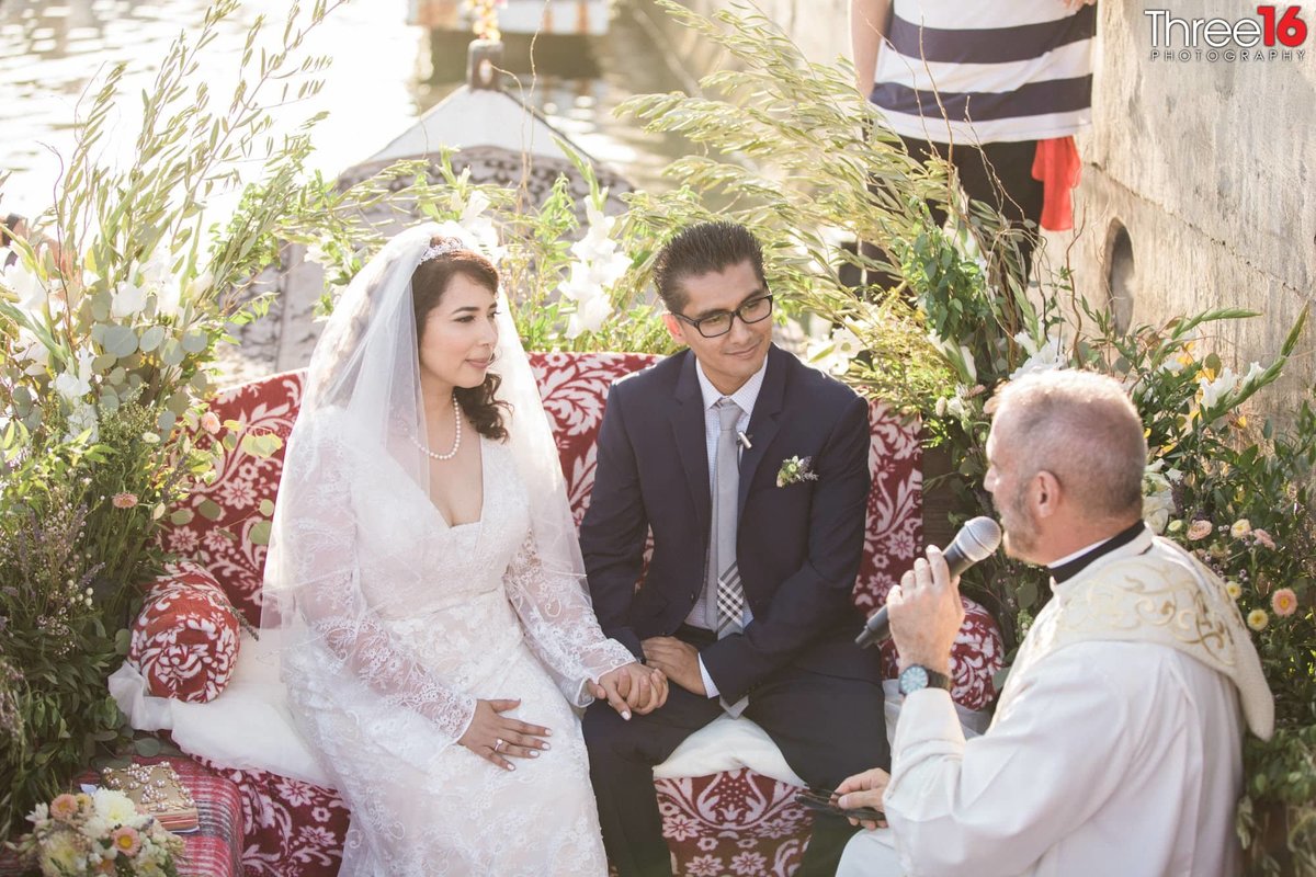 Officiant speaks into a microphone during the wedding ceremony as he, and the Bride and Groom sit during a gondola ride in Long Beach