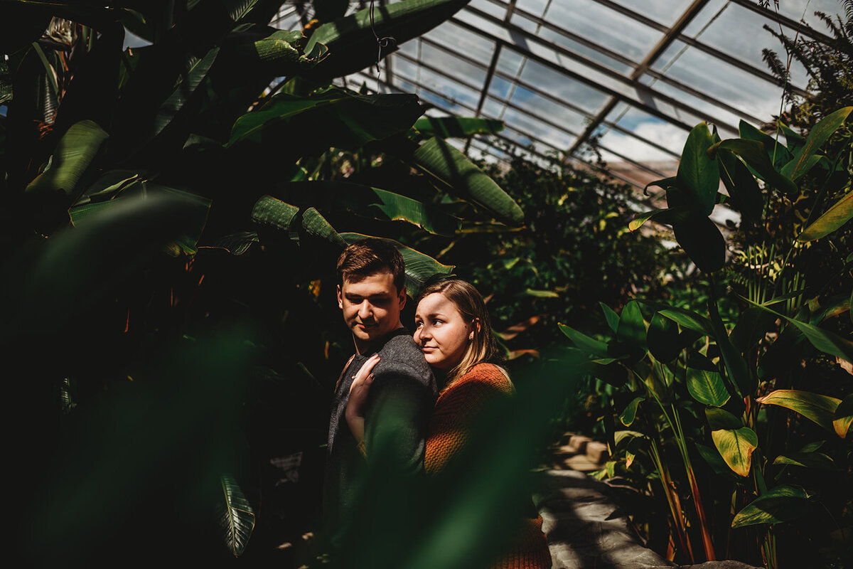Baltimore photographer captures romantic greenhouse engagement pictures with woman standing behind man and resting her cheek on his shoulder while looking up at him