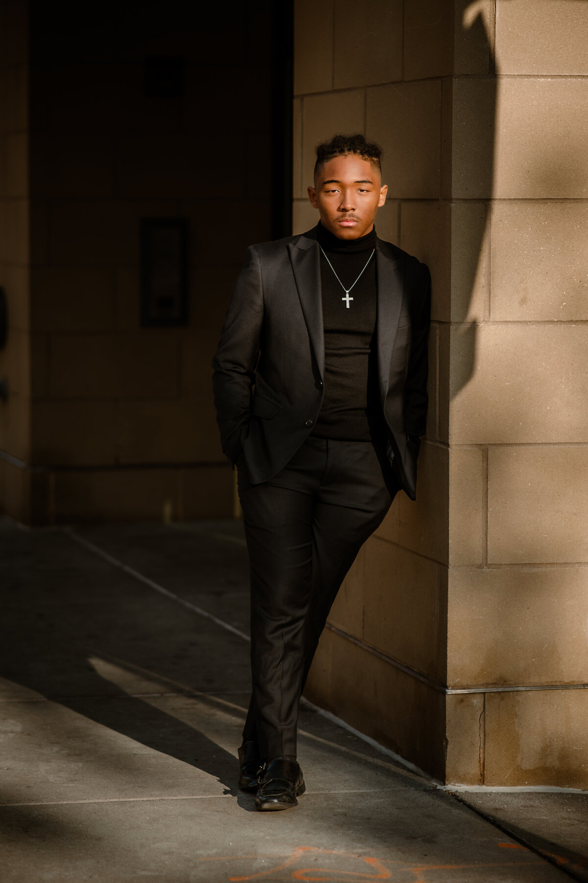 Young black man dressed in an all black suit at sunset leaning against a city building in St. Paul