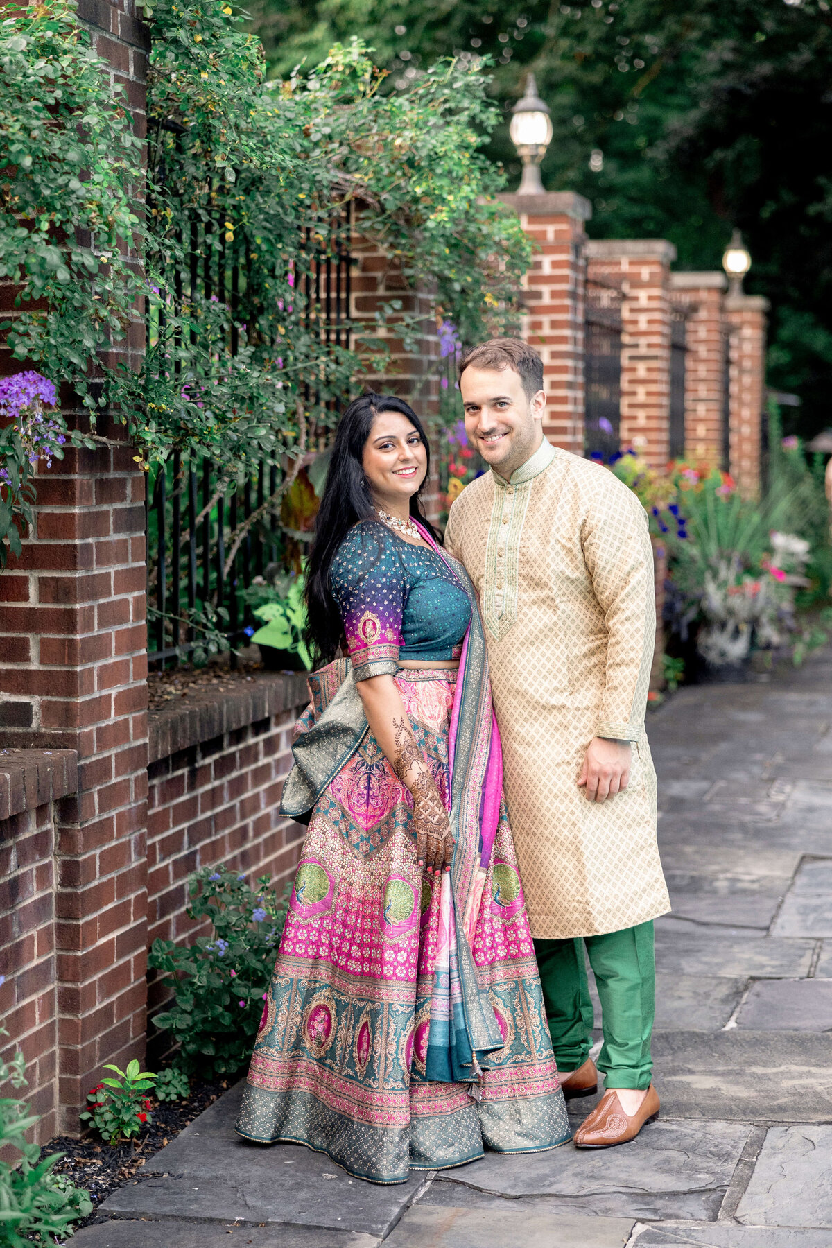 A couple stands together outdoors on a stone pathway lined with brick walls and greenery. The woman is wearing a colorful traditional dress and has long dark hair. The man is dressed in a cream and green traditional outfit. Both are smiling at the camera.