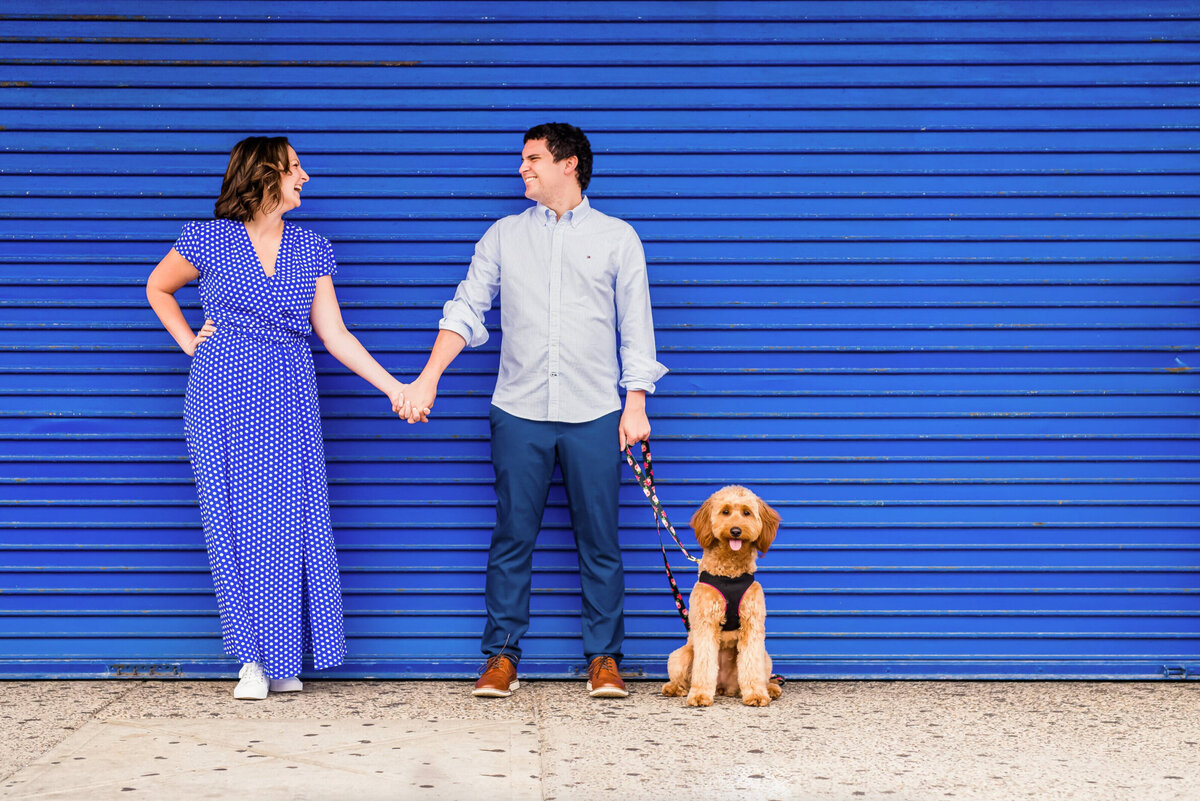 Man and woman, both dressed in blue standing in front of a blue wall with their dog on Jenkinson's boardwalk in New Jersey