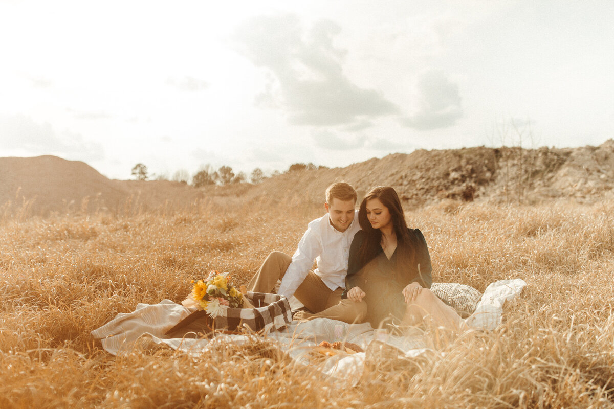 A couple is eating a charcuterie board while sitting on a picnic blanket in a field at sunset.