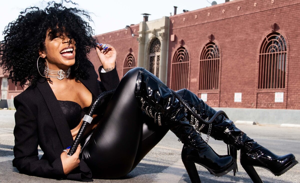 Women  music portrait Exxxoticc laughing sitting  wearing black leather outfit  brick building backdrop
