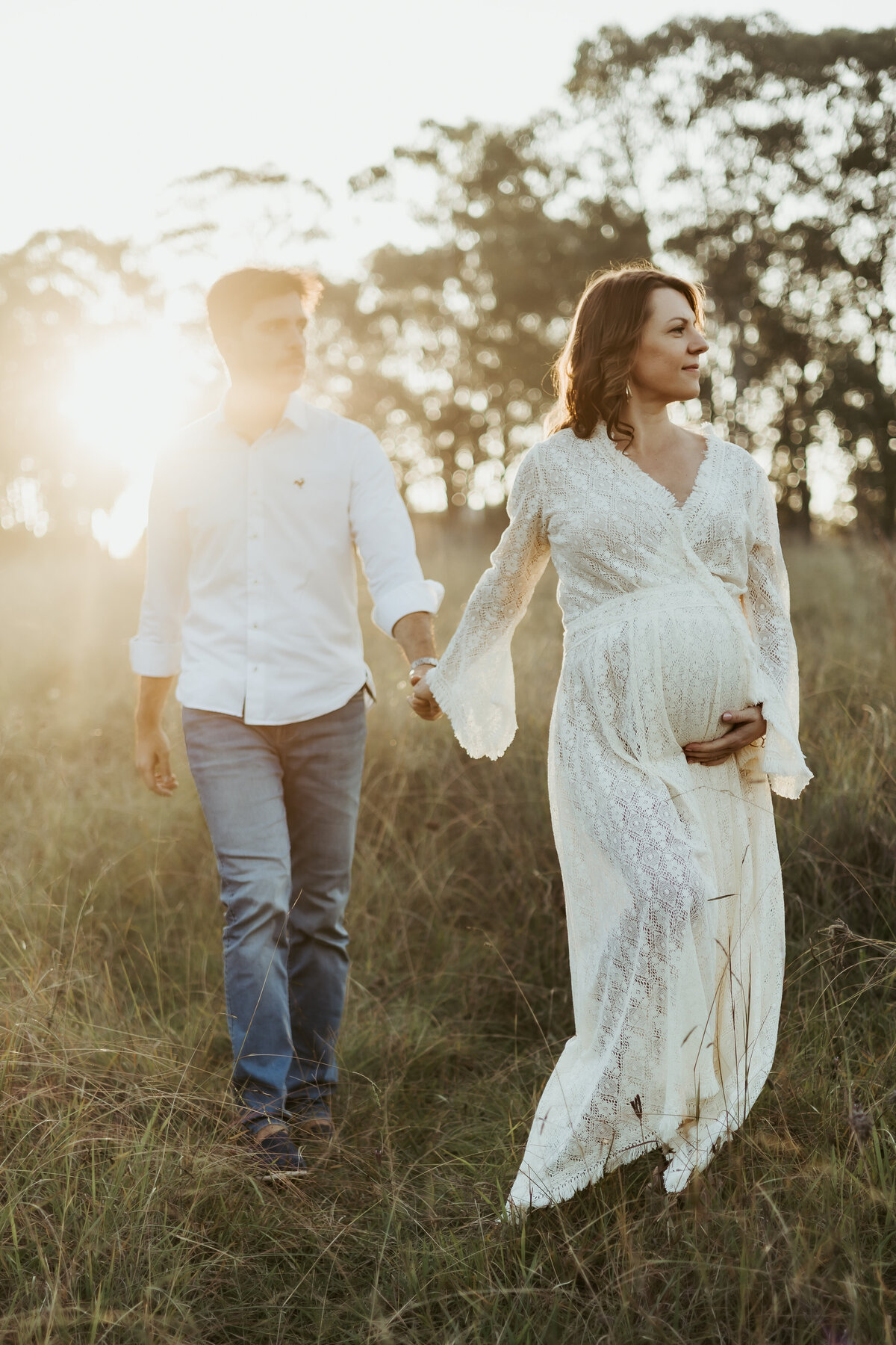 Pregnant wife in white lace robe holding her belly while she holds her husband's hand and they walk  through a field at sunset