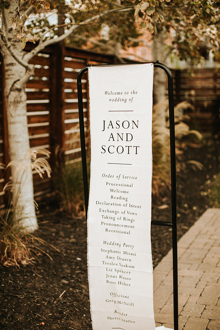 White wedding banner with black font hanging on black metal stands in the outdoors.