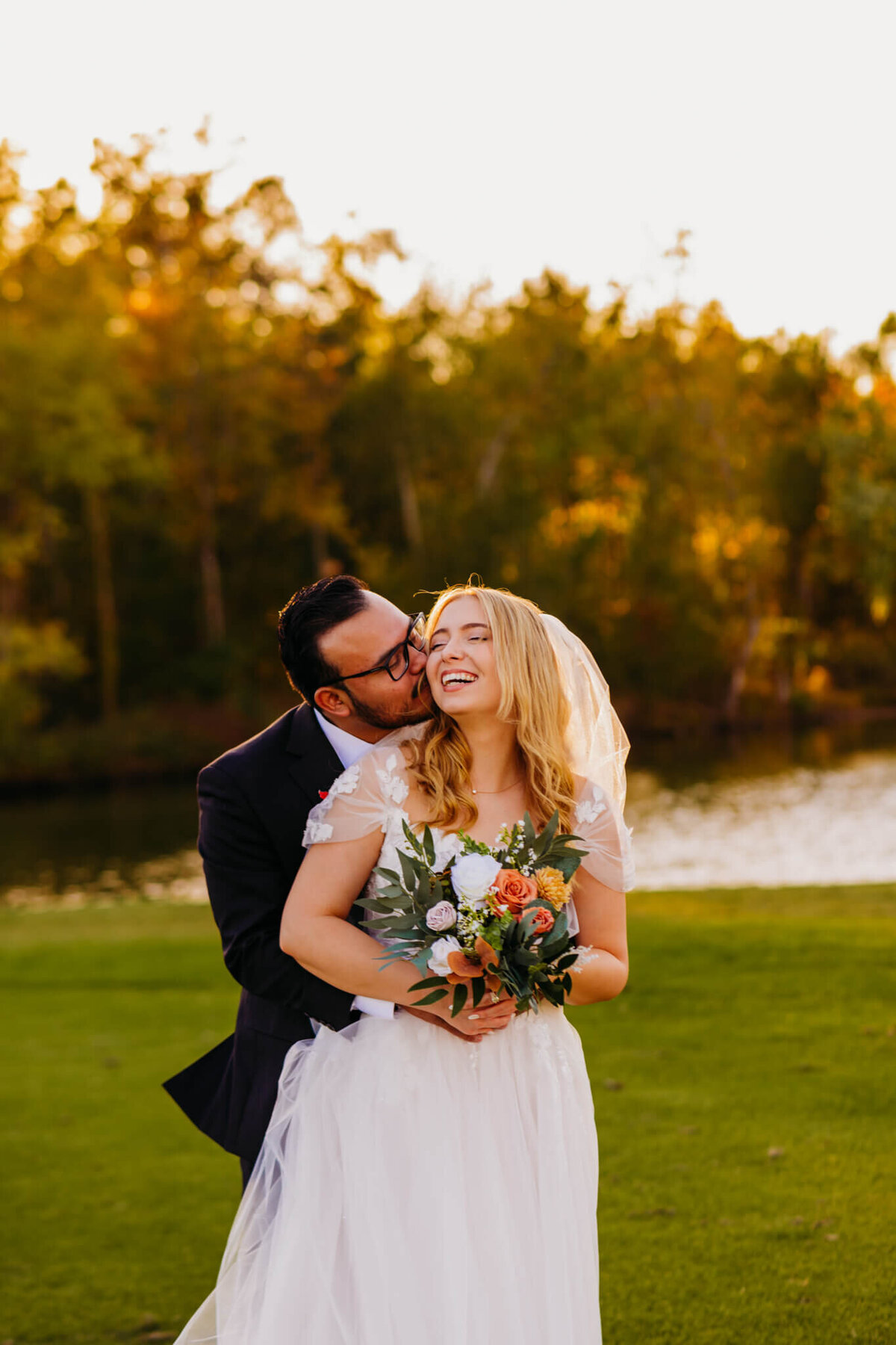 photo of a bride laughing while the groom hugs her from behind and kisses her cheek