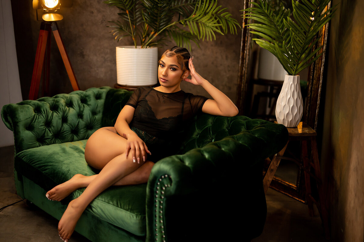 A woman perches on an emerald green, velvet sofa wearing a sheer, black body suit in our Waukesha photo studio.