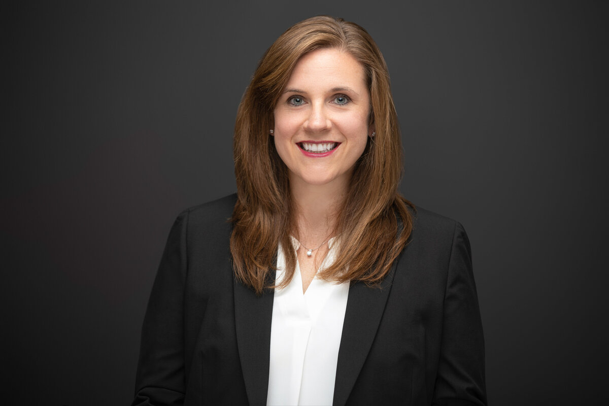 An executive businesswoman with long brown hair wearing a navy black blazer poses for a professional headshot on a black background for Janel Lee Photography studios Cincinnati Ohio
