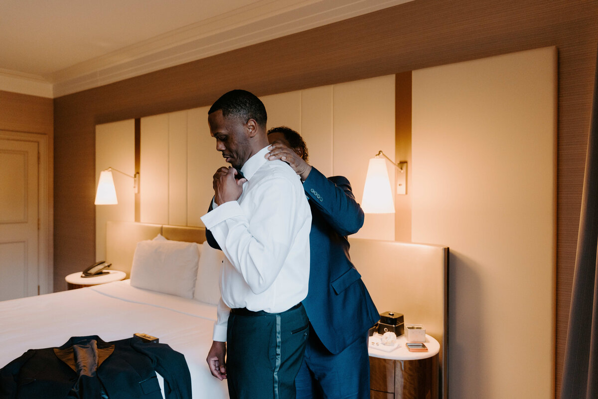 A groom in a white shirt is helped with his tie by another person in a cozy hotel room. Warm lighting creates an intimate and calm atmosphere.