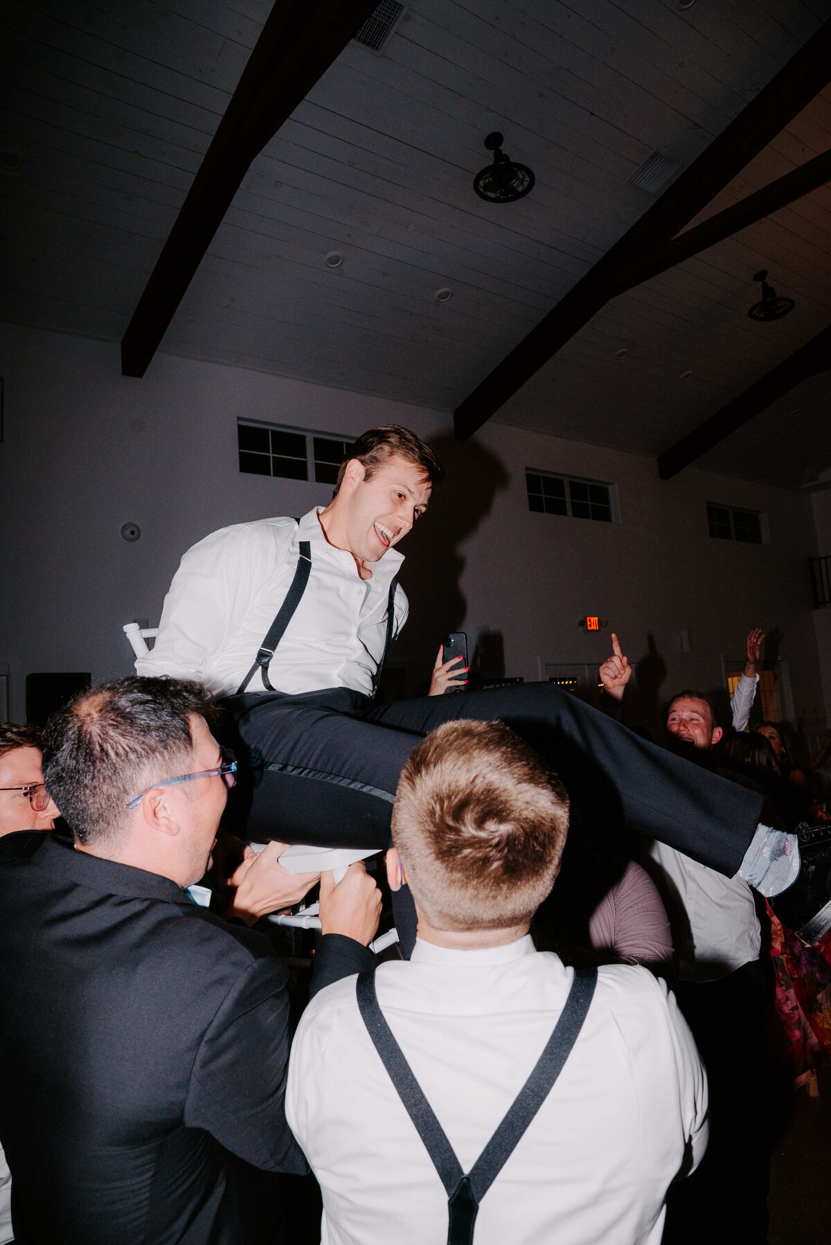 A joyful man being lifted in the air by a group of friends at a lively indoor party. They are all dressed in formal attire, conveying a celebratory mood.