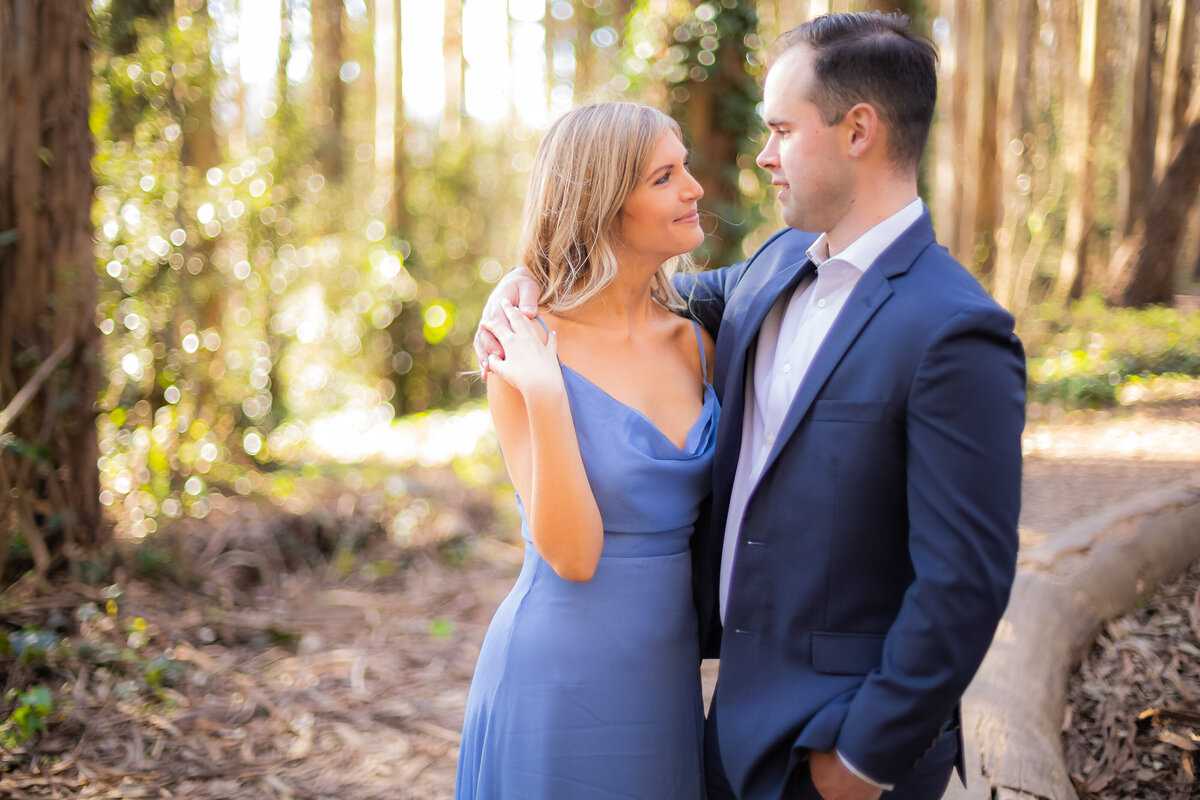 Engaged couple looks at each other smiling in the woods. The girl wears lavender and the man wears a blue suit. He has his arm around her neck. Photo by wedding photographer, philippe studio pro.