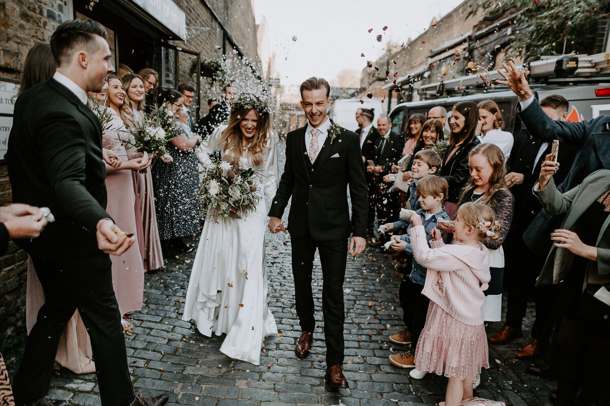 A couple walk down a confetti line at Clapton Country Club in London. The alternative wedding venue has some awesome spots for couples portraits and a cobbled road.