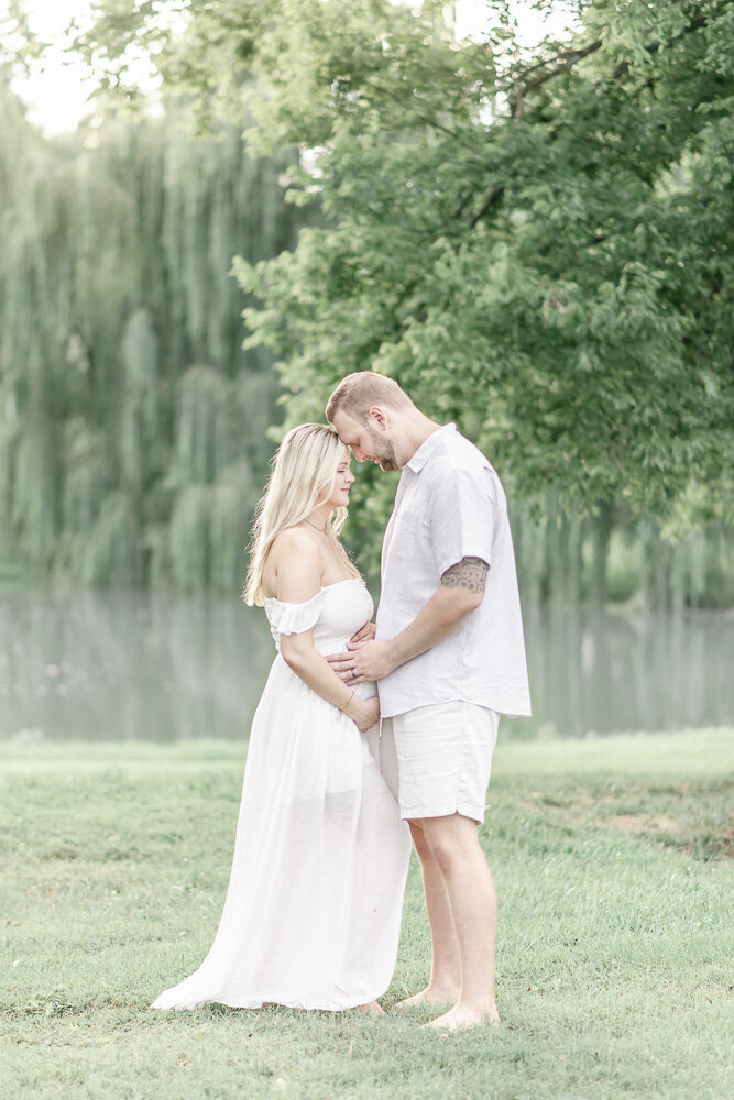 Pregnant couple hug standing in front of a willow tree