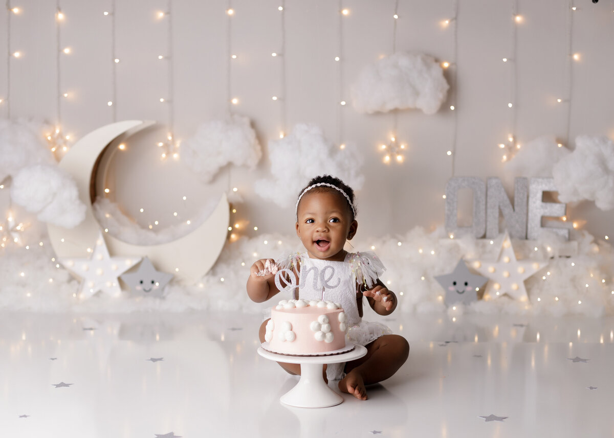 Moon and Stars theme cake smash.  Baby is sitting behind a peach cake smiling at the camera.  In the background there are faux clouds, light up stars and a wooden moon with fluffy clouds along the bottom.