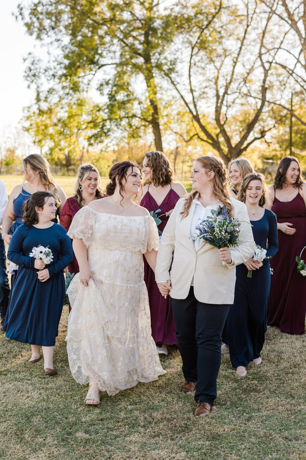 Two brides walking through nature with their wedding party after their wedding ceremony in Dallas, Texas. The bride on the right is wearing a cream jacket, dark dress pants, and is holding a bouquet. The bride on the left is wearing an intricate, flowing, white dress. The wedding party is wearing dresses of either blue or burgundy and many of them are carrying small bouquets.