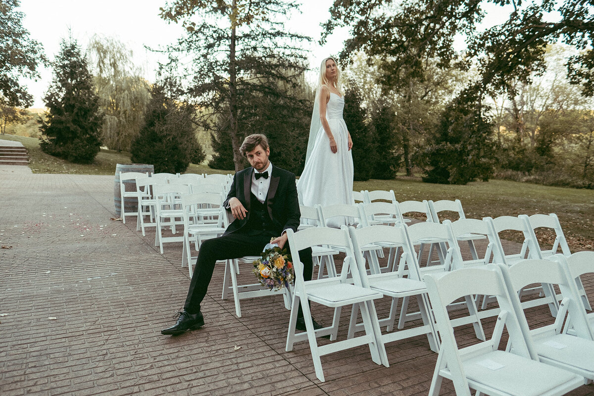 A groom sitting on a chair with a bouquet looks to the side, while a bride in a white dress stands on a chair behind him, outdoors at an Iowa wedding venue.