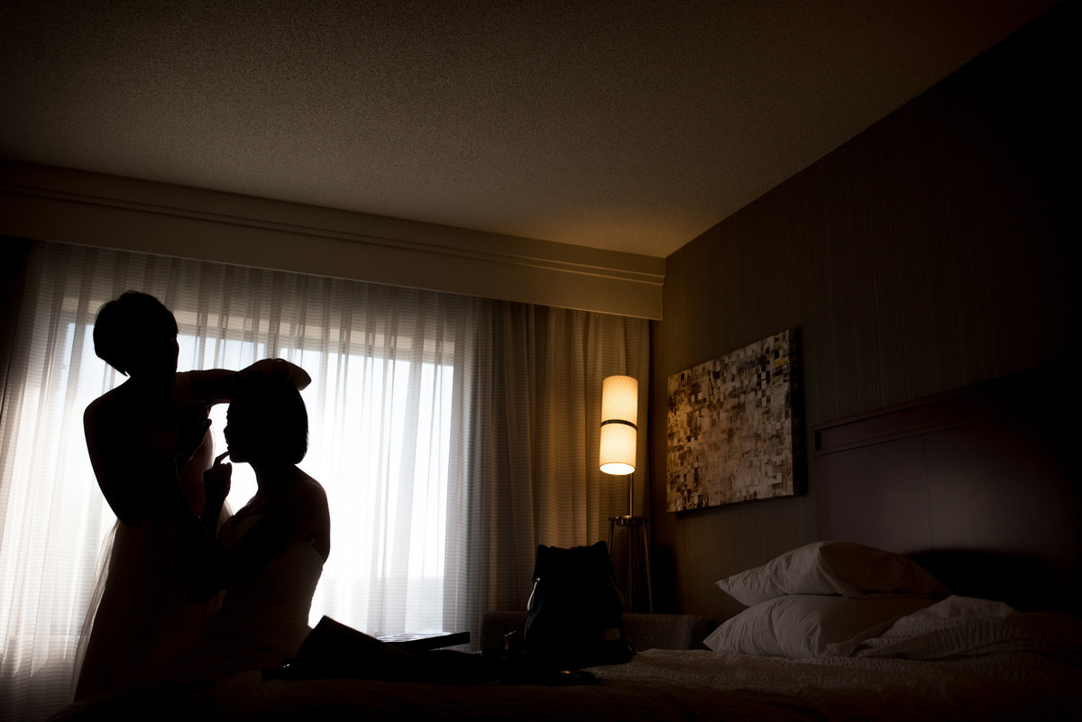 The bride is silhouetted against the window of a hotel room while getting her hair and makeup done