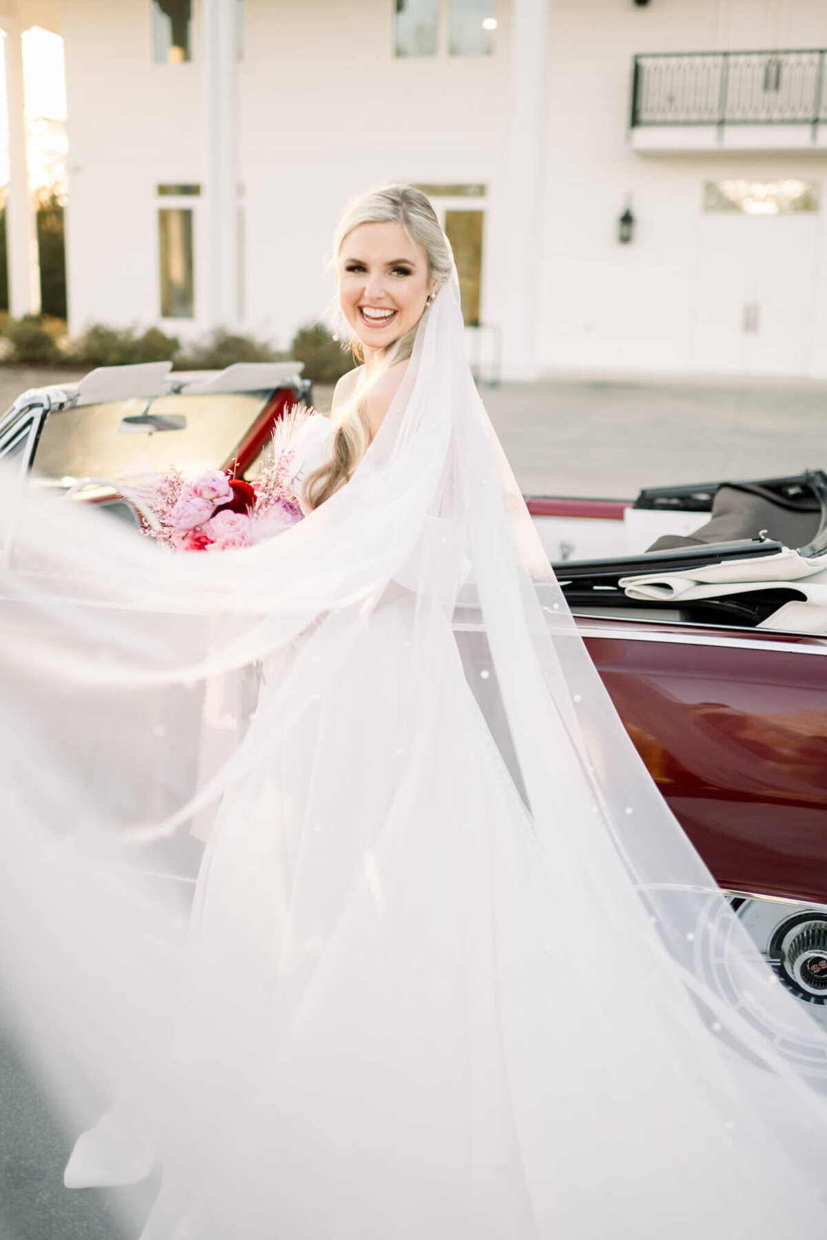 bride smiles at camera in front of legacy acres wedding venue and red vintage car with long veil  draped in front of her