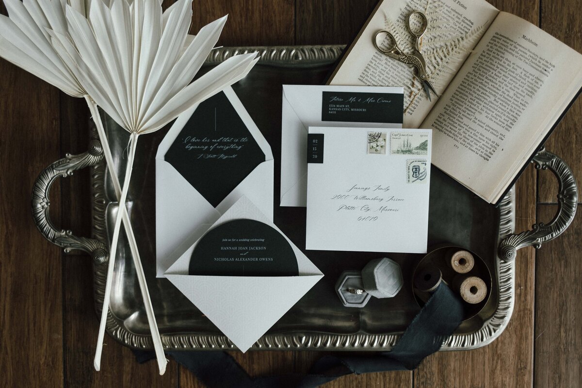 Mixed white wedding stationery with cursive fonts set on a silver tray with a book, twine and scissors on a wooden floor.