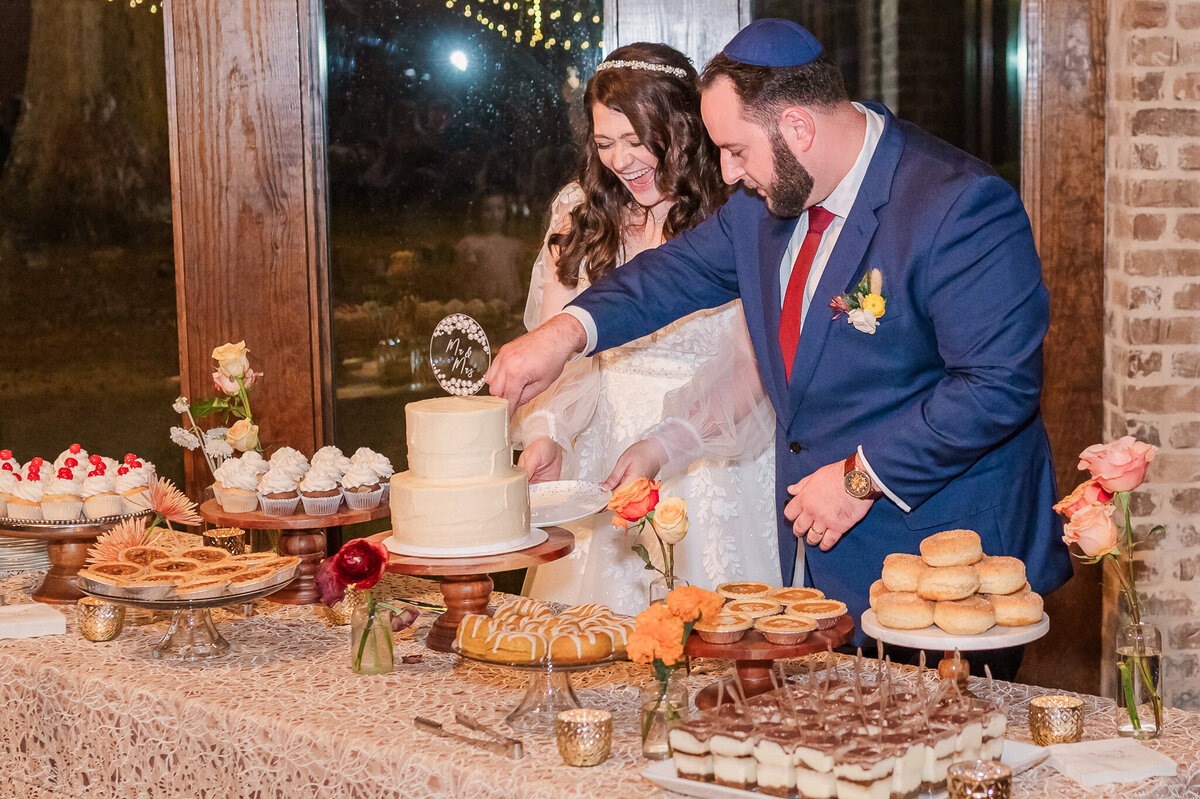 A happy bride and groom cutting a wedding cake surrounded by desserts on their wedding day enjoying their Raleigh wedding photography session by JoLynn Photography