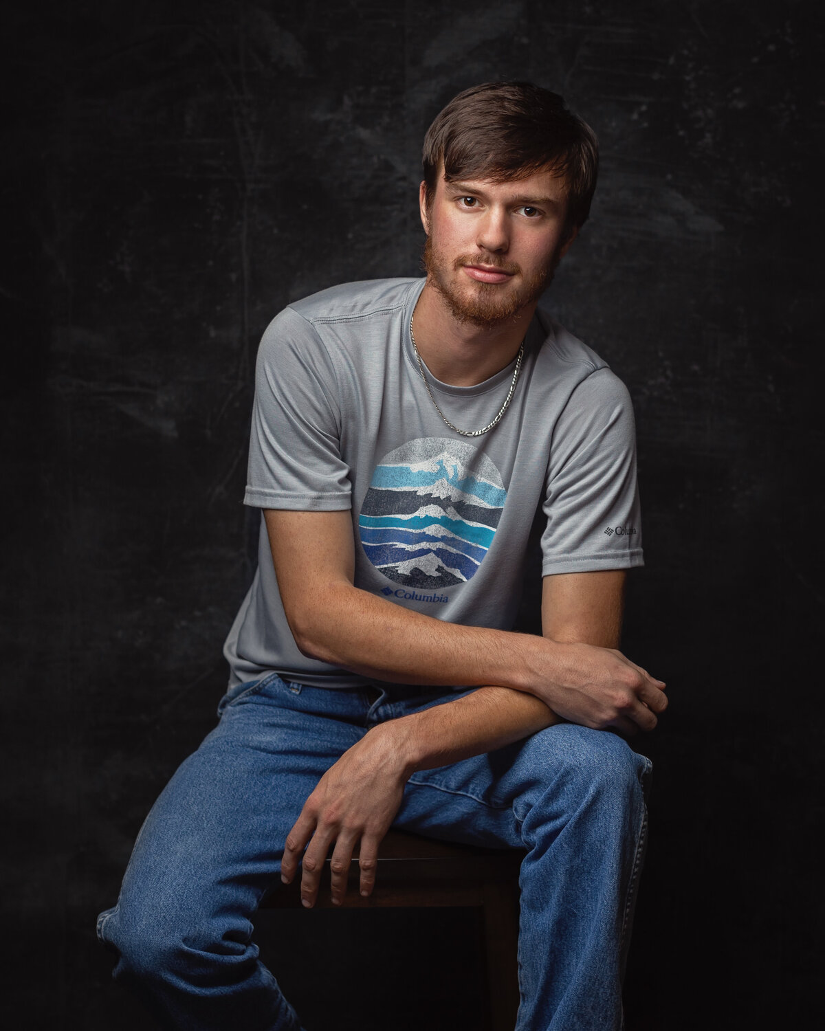 man in blue jeans and a shirt seated in a studio with both hands crossed  leaning on left leg