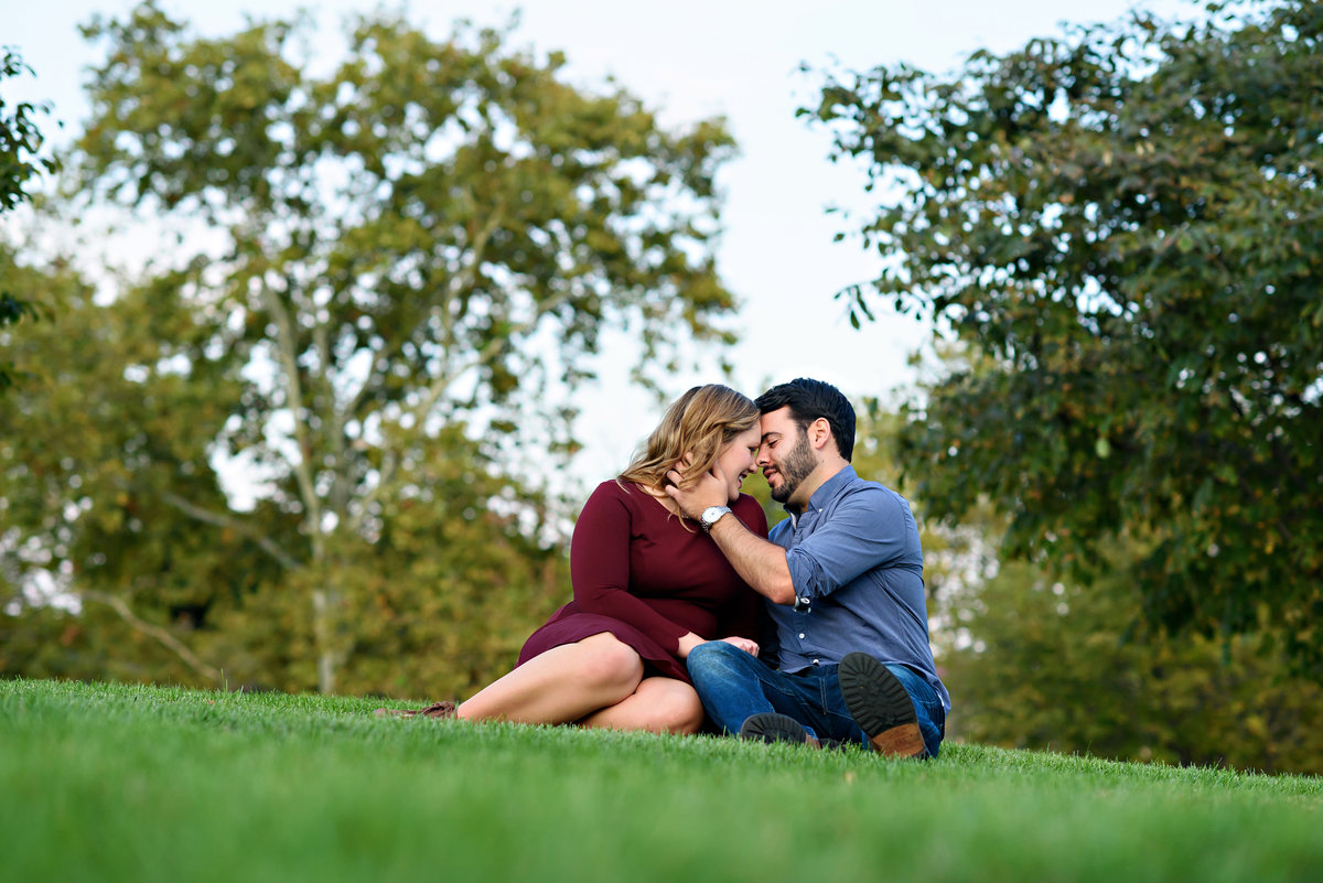 A man brings his fiance in for a kiss while sitting on the grass at the Philadelphia Museum of Art.